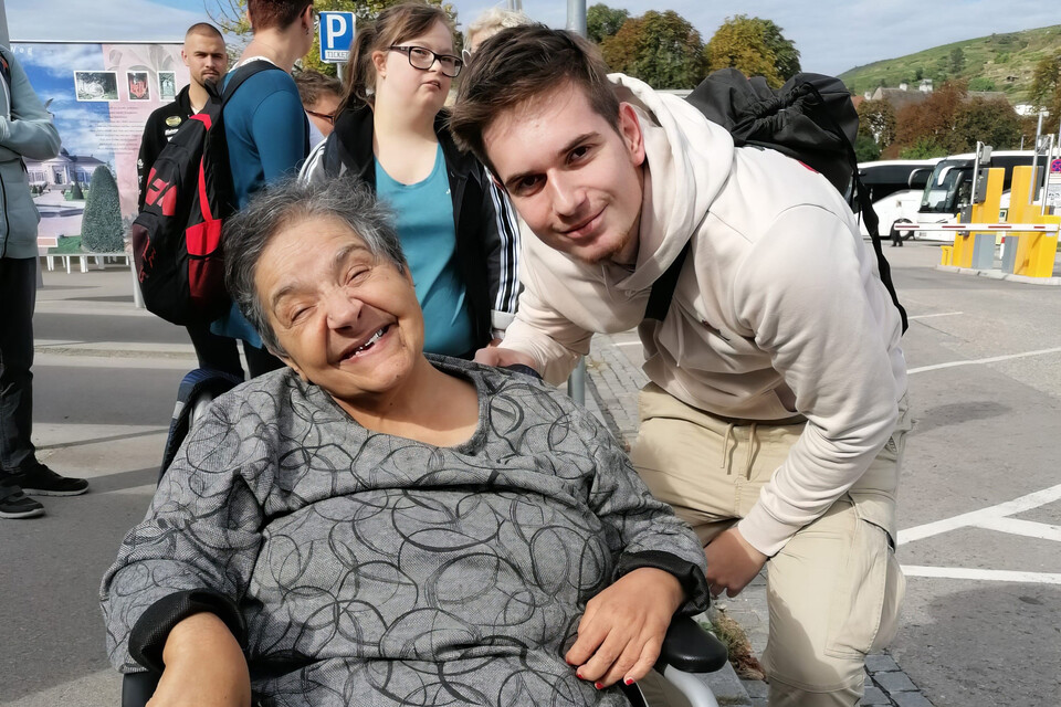 Ein Zivildiener posiert auf einem Ausflug gemeinsam mit einer Bewohnerin, die im Rollstuhl sitzt.