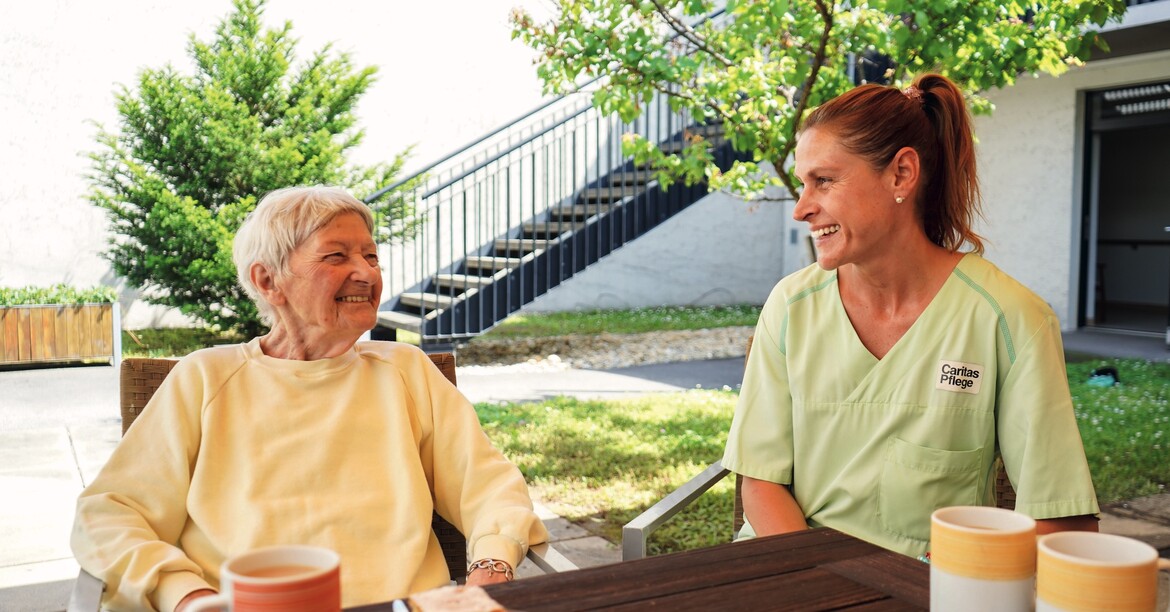 Eine Pflegerin sitzt lachend mit einer Bewohnerin bei einer Jause im Garten. 