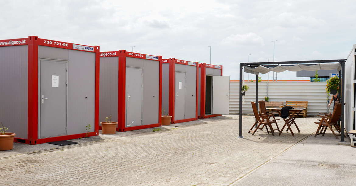 Mehrere rot-graue Container im Freien, gegenüber ein überdachter Sitzbereich.