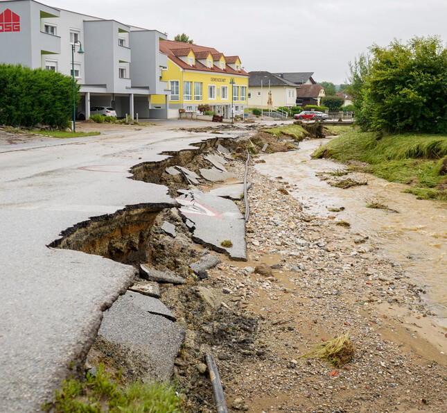 Eine von einem Hochwasser zerstörte Straße ist aufgebrochen, während ein überfluteter Bach neben der Straße fließt. Im Hintergrund sind Wohnhäuser zu sehen.