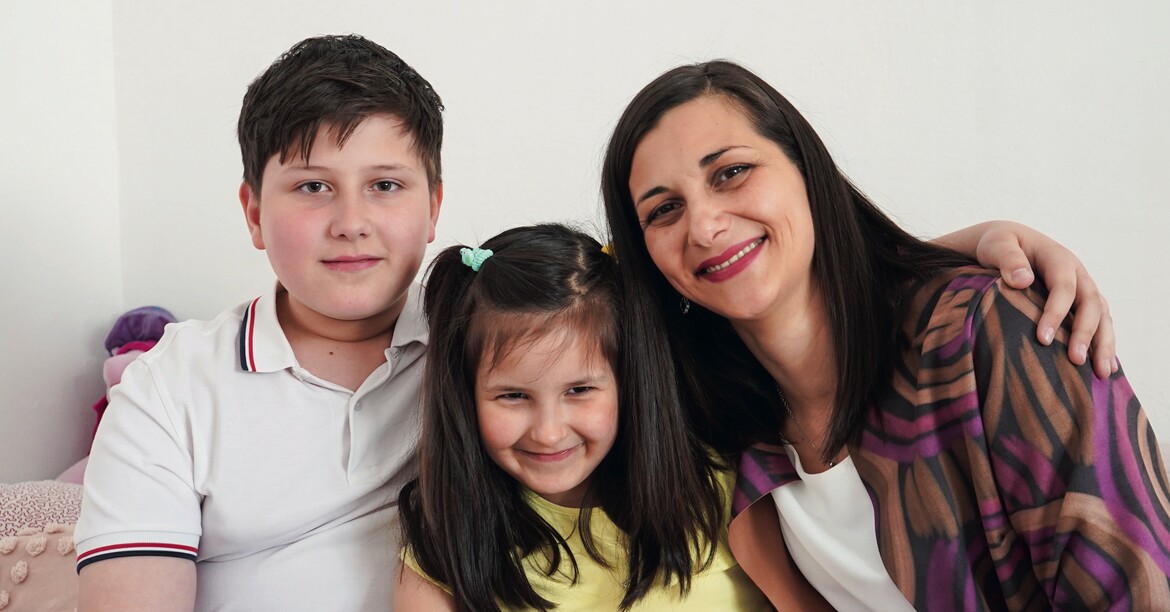 Eine Mutter mit ihren zwei Kindern sitzt lächelnd auf einem Sofa. 