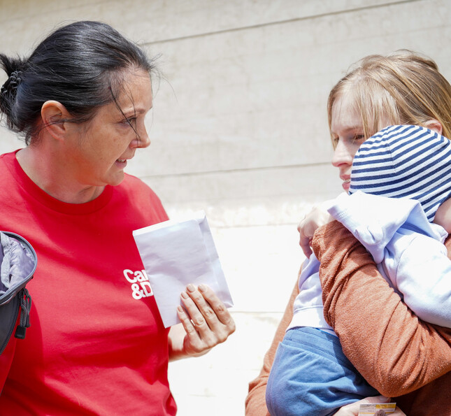Caritas Direktorin Melanie Balaskovics redet mit einer vom Hochwasser betroffenen Person, welche ein Baby auf dem Arm hält. Die Frau bekommt ein Kuvert überreicht.