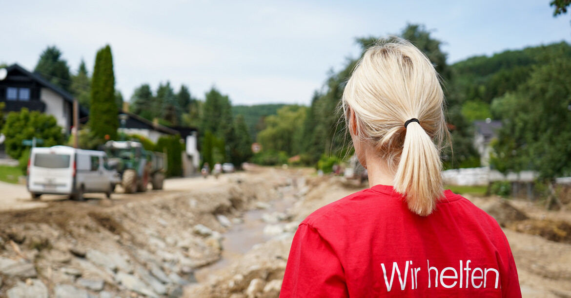 Eine Caritas Mitarbeiterin steht im Vordergrund mit dem Rücken zum Bild und blickt zu den Aufräumarbeiten nach dem Hochwasser.