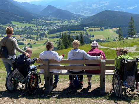 Haus Vitus Bewohner*innen und Betreuer*innen sitzen auf einer Bank und genießen den Ausblick. Zu sehen ist eine Grünfläche mit Bergen im Hintergrund.