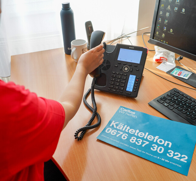 Eine Frau legt gerade den Hörer auf in einem Büro. Auf dem Tisch liegt ein beklaue Flyer mit Telefonnummer drauf. 
