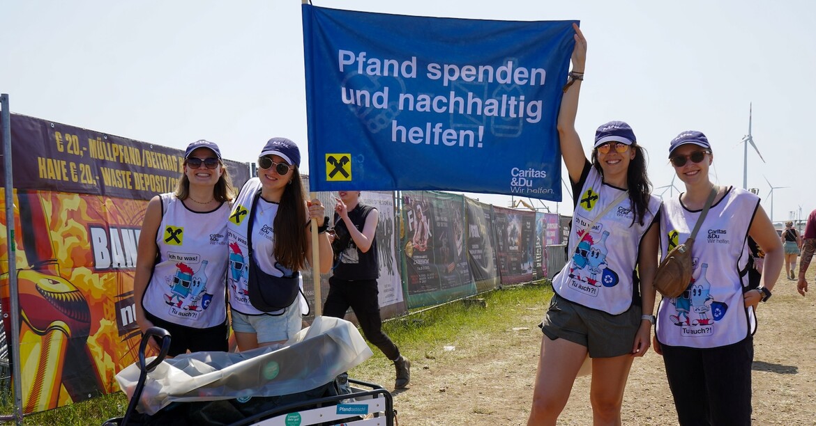 Vier lächelnde Frauen mit Caritas Weste halten eine blaue Fahne in die höhe beim Pfandsammeln am Festival. 