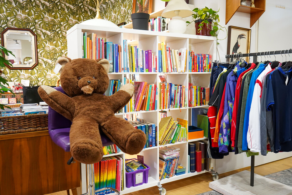 Ein Bereich des carla Shops im Burgenland mit einem großen braunen Plüschbären, einem Regal voller bunter Bücher und einem Kleiderständer mit Secondhand-Kleidung.