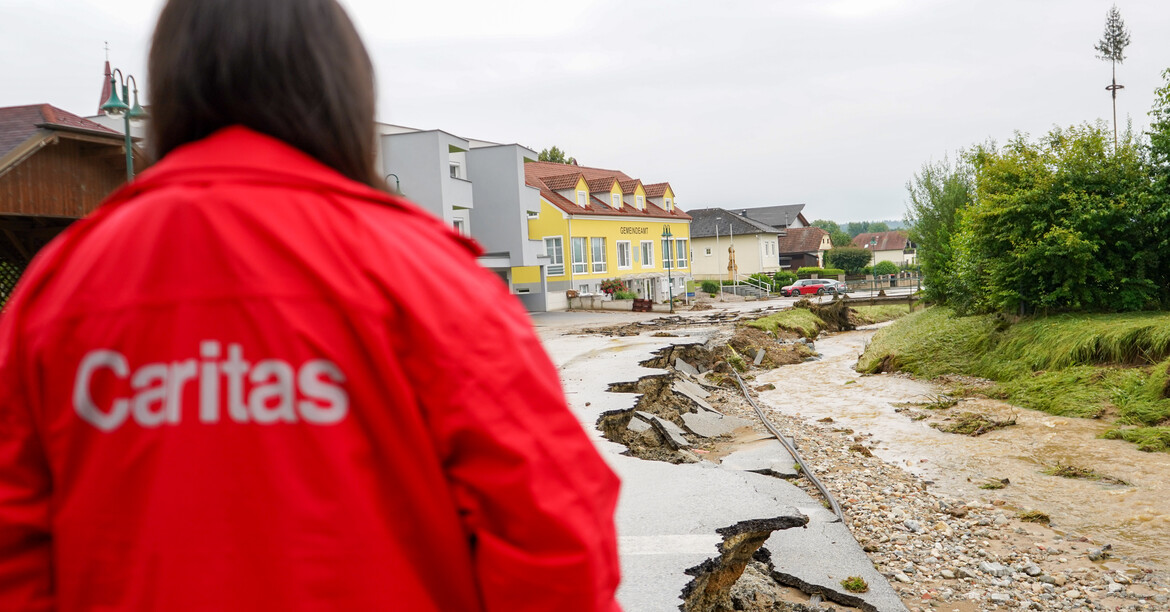 Eine Person in einer roten Caritas Jacke steht vor einer durch Hochwasser stark beschädigten Straße, deren Oberfläche aufgebrochen ist, während ein überfluteter Bach neben der Straße vorbeifließt.