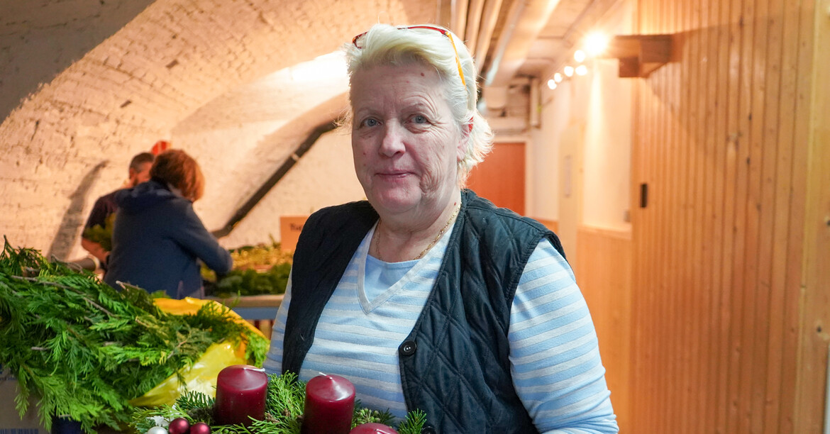 Eine ältere Frau schaut lächelnd in die Kamera und hat einen Adventkranz in der Hand.