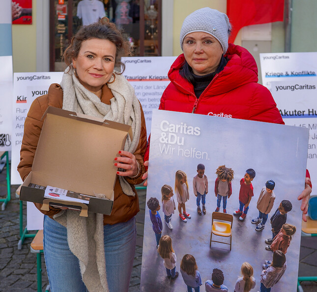 Klassenvorständin Martina Stolavetz und Caritas Direktorin Melanie Balaskovics stehen vor den Stühlen mit Schildern.