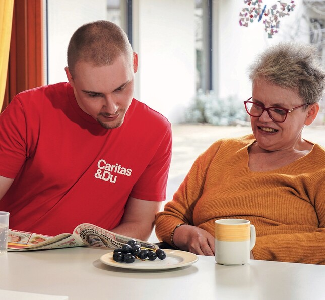 Ein Pflegelehrling sitz mit einer Bewohnerin bei einer Jause. 